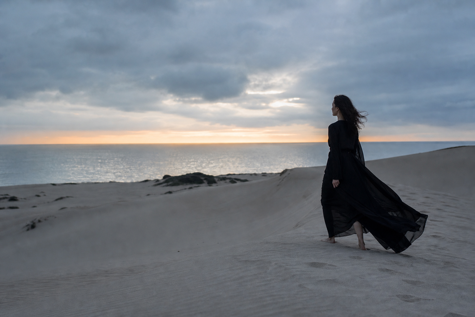 Woman in a black dress walking across coastal dunes at dusk